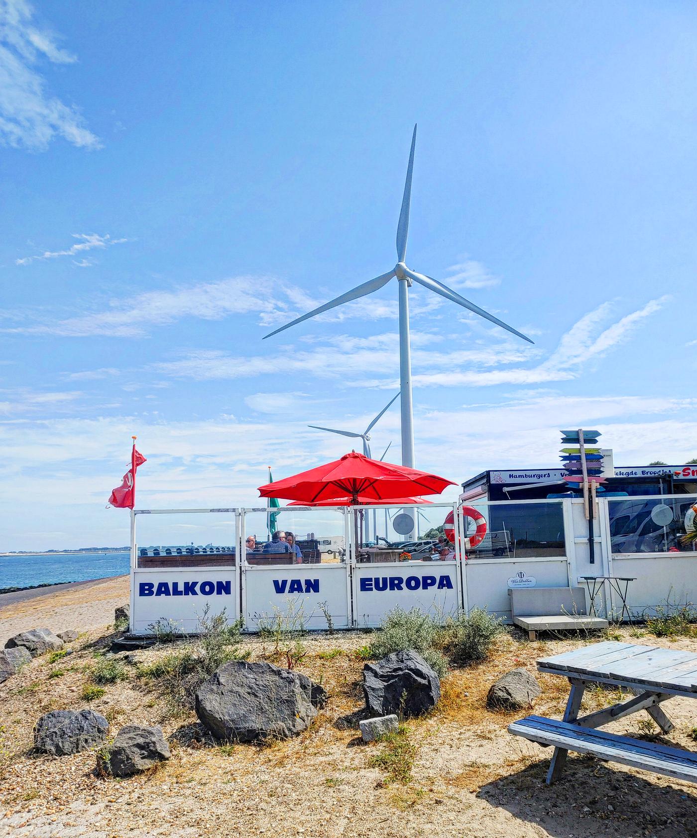 Het bordje "Balkon van Europa" op het terrashek, met een grote windmolen op de achtergrond