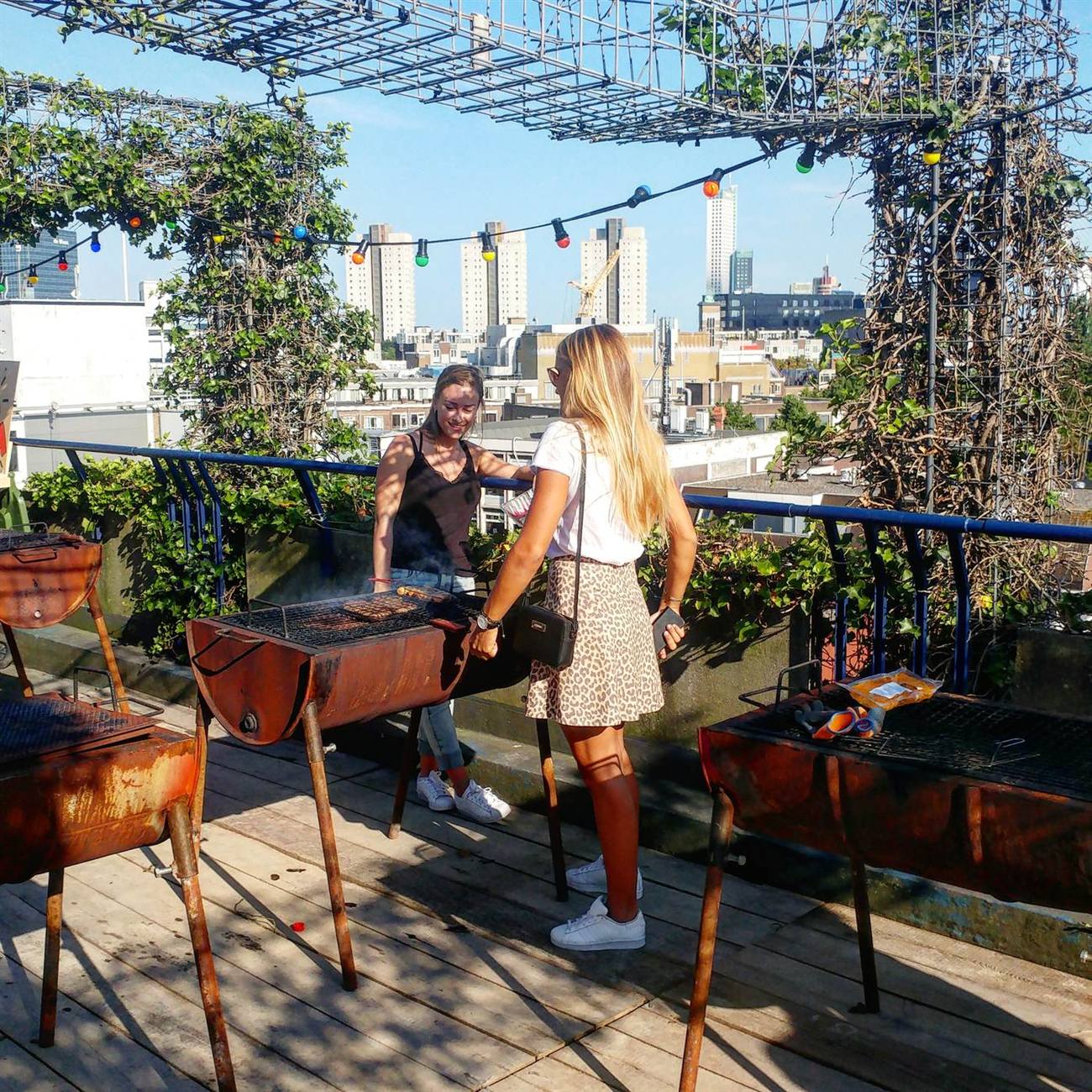 Twee vrouwen staan bij een roestige vatenbarbecue in het gouden avondlicht, met de Rotterdamse skyline op de achtergrond