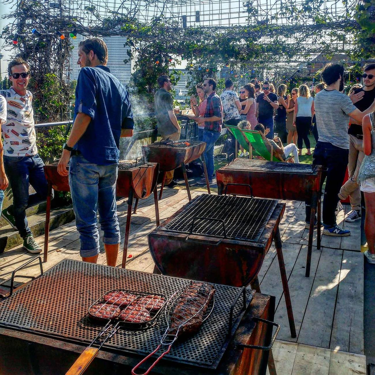 Bezoekers grillen vlees op de vatenbarbecues, met zonlicht en de stad op de achtergrond, zomer 2017
