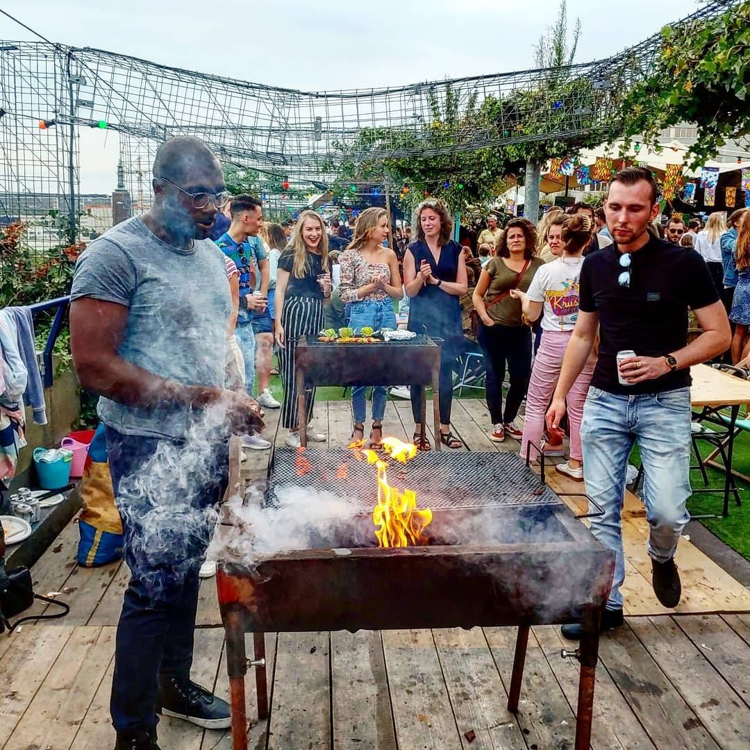 Een man staat in de rook bij een brandende bbq op het dak, terwijl bezoekers eromheen kletsen met een drankje in de hand