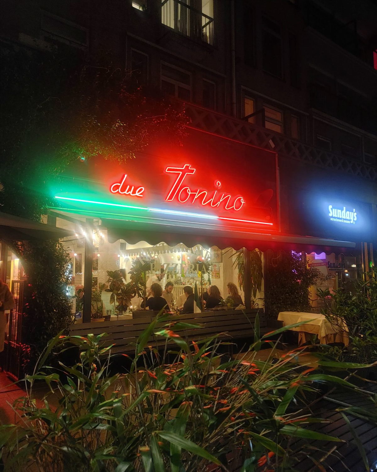 De gevel van Due Tonino in de avond, met neonletters in de kleuren van de Italiaanse vlag en gasten aan tafel achter het raam.