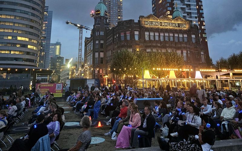 Publiek van openluchtbioscoop Pleinbios op de Wilhelminapier, met het Holland Amerika Lijn-gebouw van Hotel New York op de achtergrond