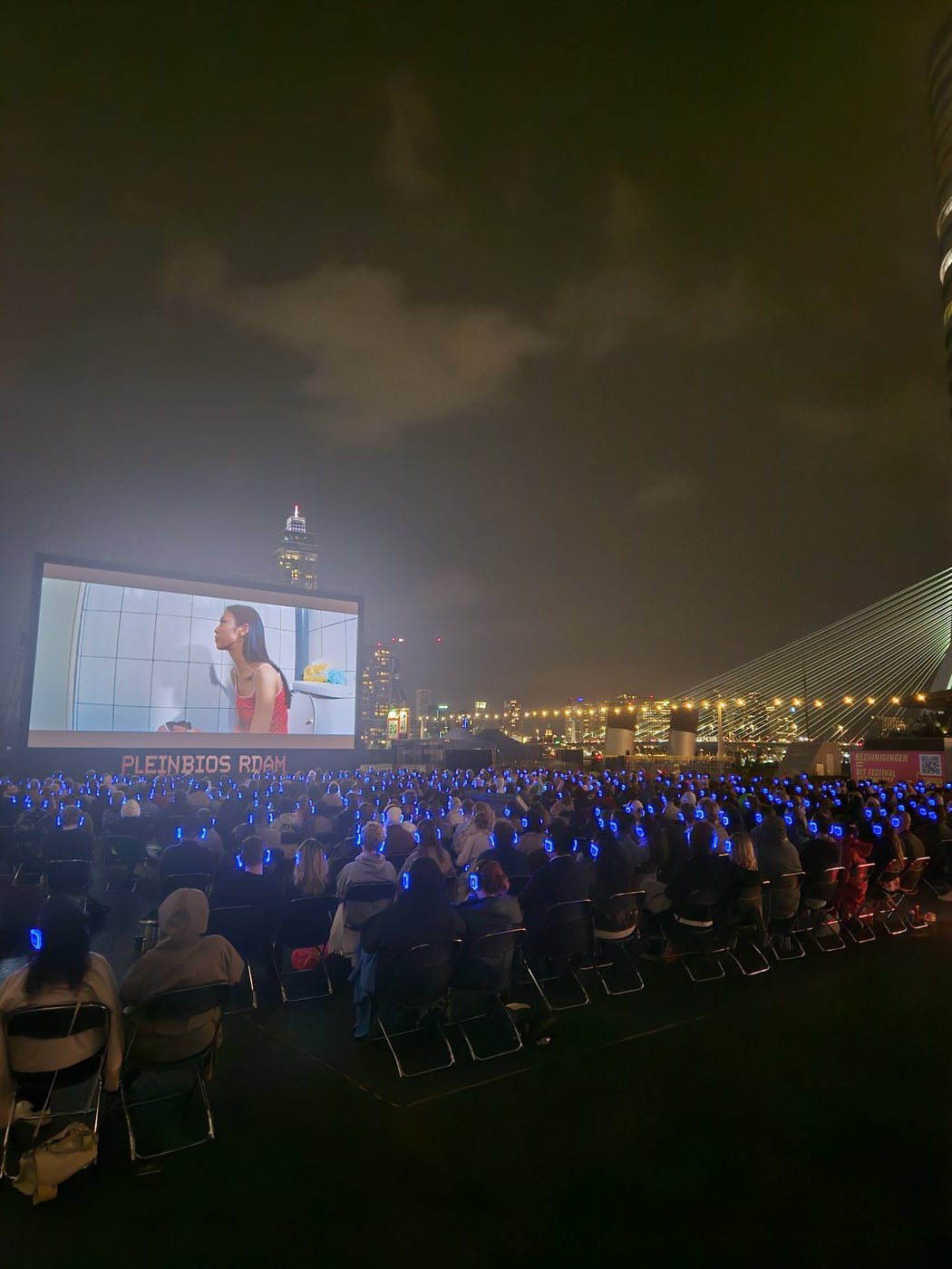 Honderden blauw oplichtende koptelefoons in het donker bij Pleinbios, met de Erasmusbrug en de skyline op de achtergrond.