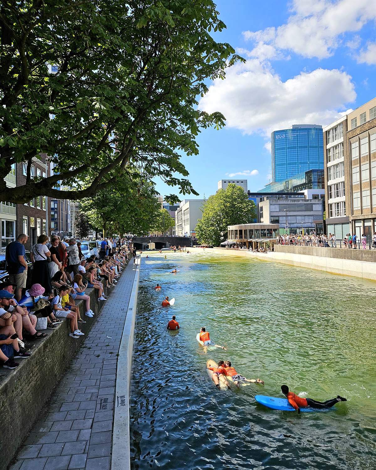 Toeschouwers kijken vanaf de kade naar surfers in het groene water van RiF010, met de Rotterdamse hoogbouw op de achtergrond.
